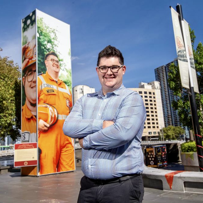 David, wearing a blue shirt and glasses, standing on the Crown Riverwalk in Melbourne, arms crossed and smiling, blue sky, in front of a big picture of himself dressed in his orange VICSES volunteer uniform.