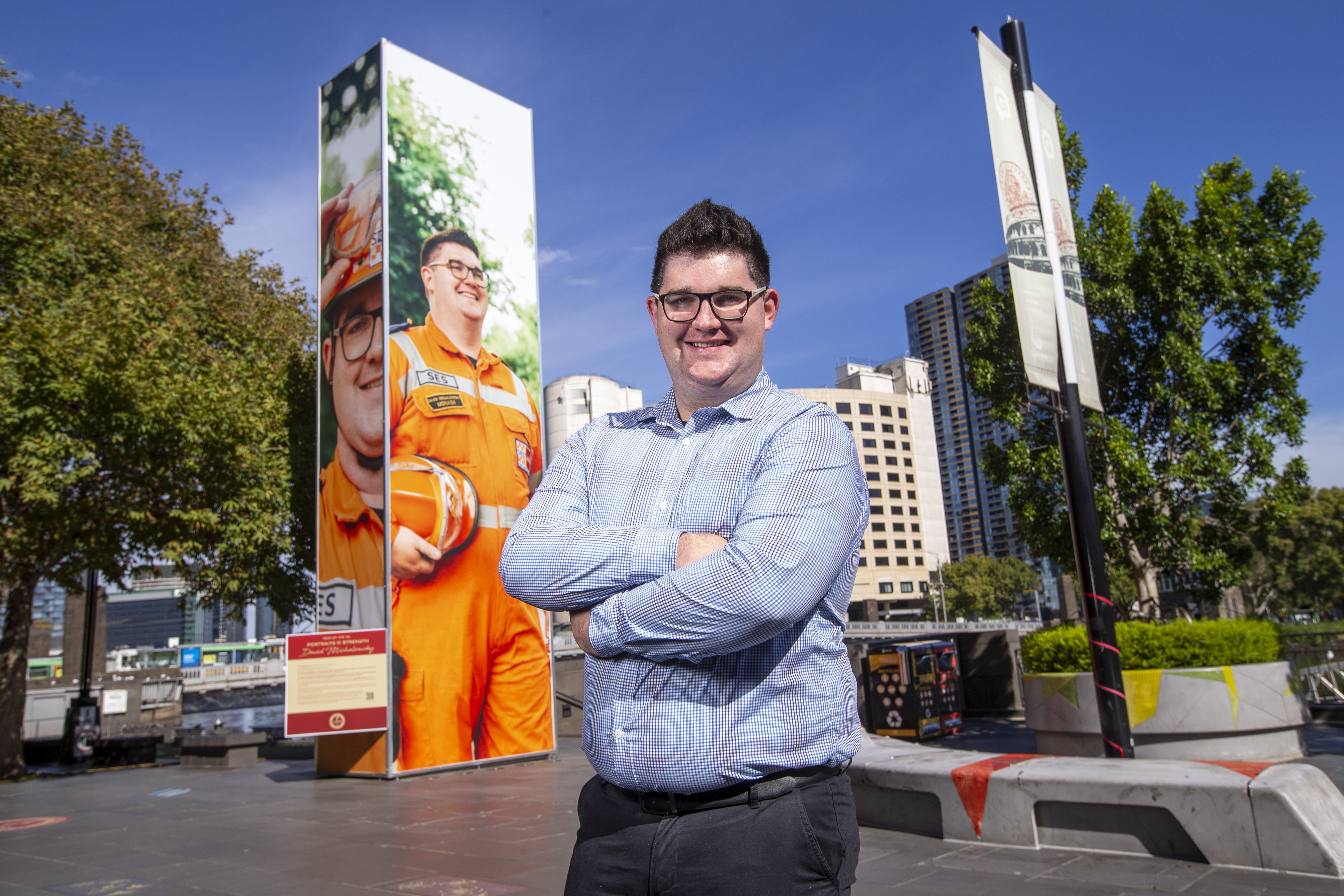David, wearing a blue shirt and glasses, standing on the Crown Riverwalk in Melbourne, arms crossed and smiling, blue sky, in front of a big picture of himself dressed in his orange VICSES volunteer uniform.