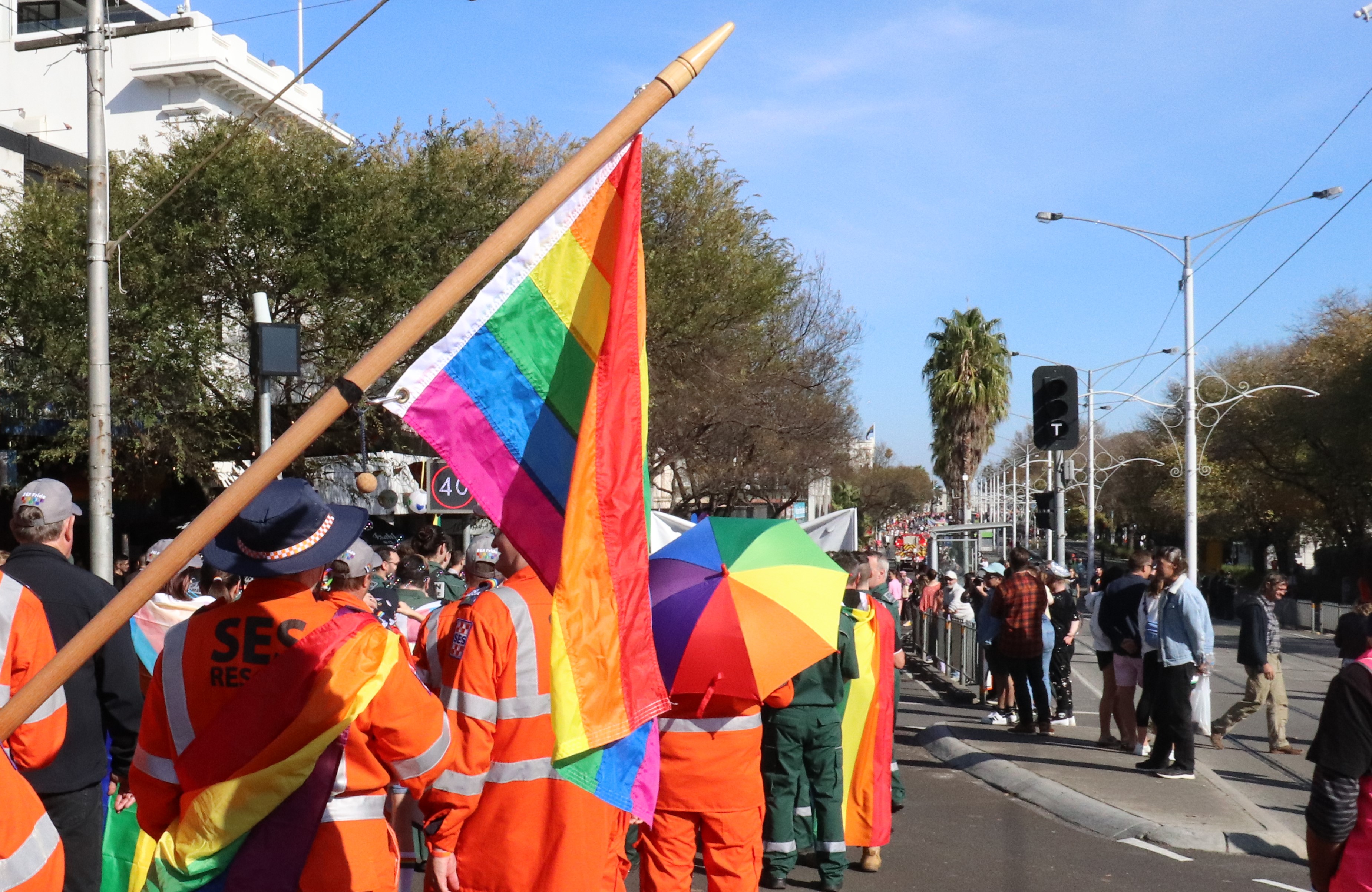 A photograph behind VICSES volunteers in orange uniforms holding rainbow flags, marching as part of Pride March. There is a blue sky above, some trees, and some people watching on from the side of the road. You cannot see anyone's faces.