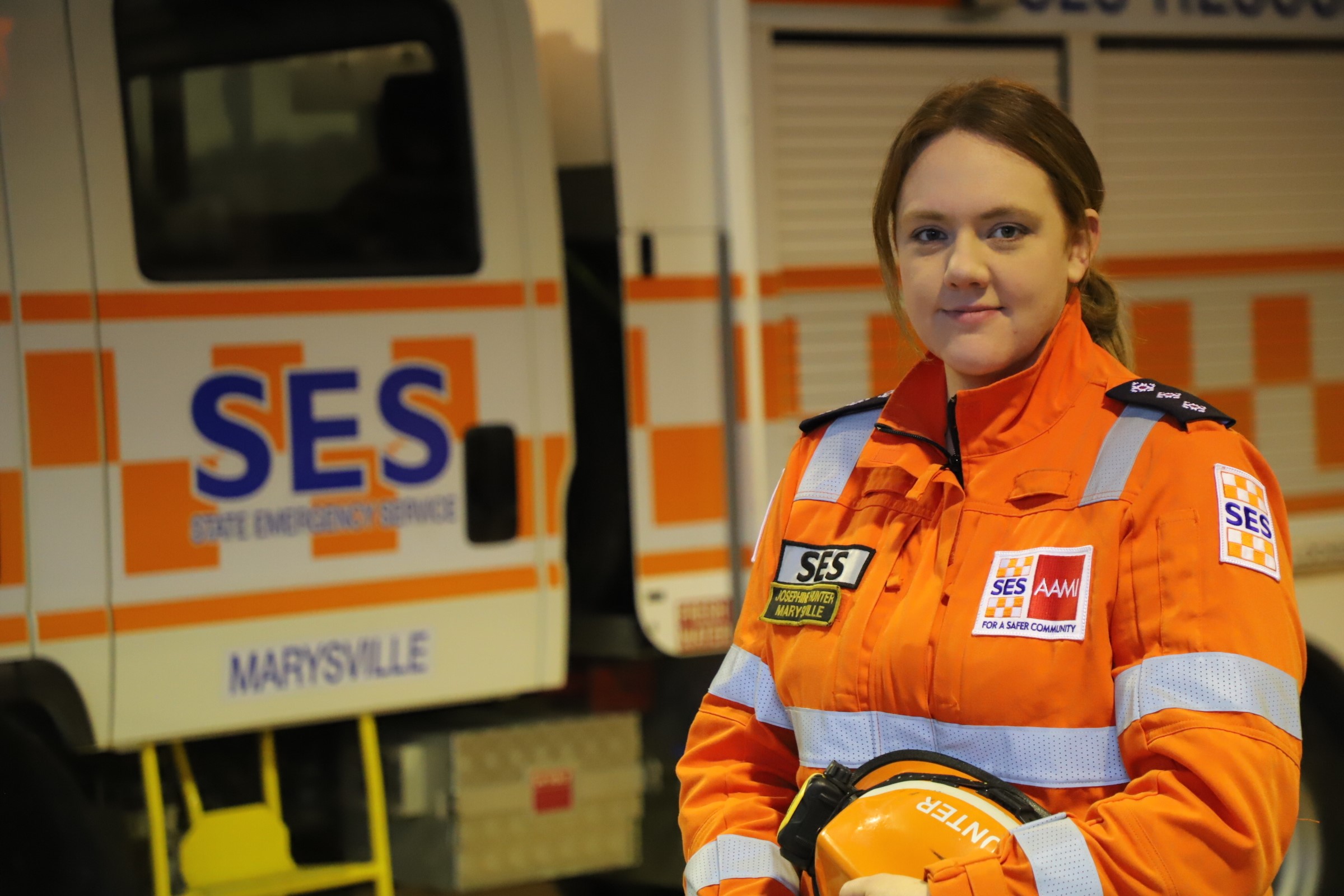 Female volunteer Jo Hunter faces camera in orange SES uniform with an SES truck in the background